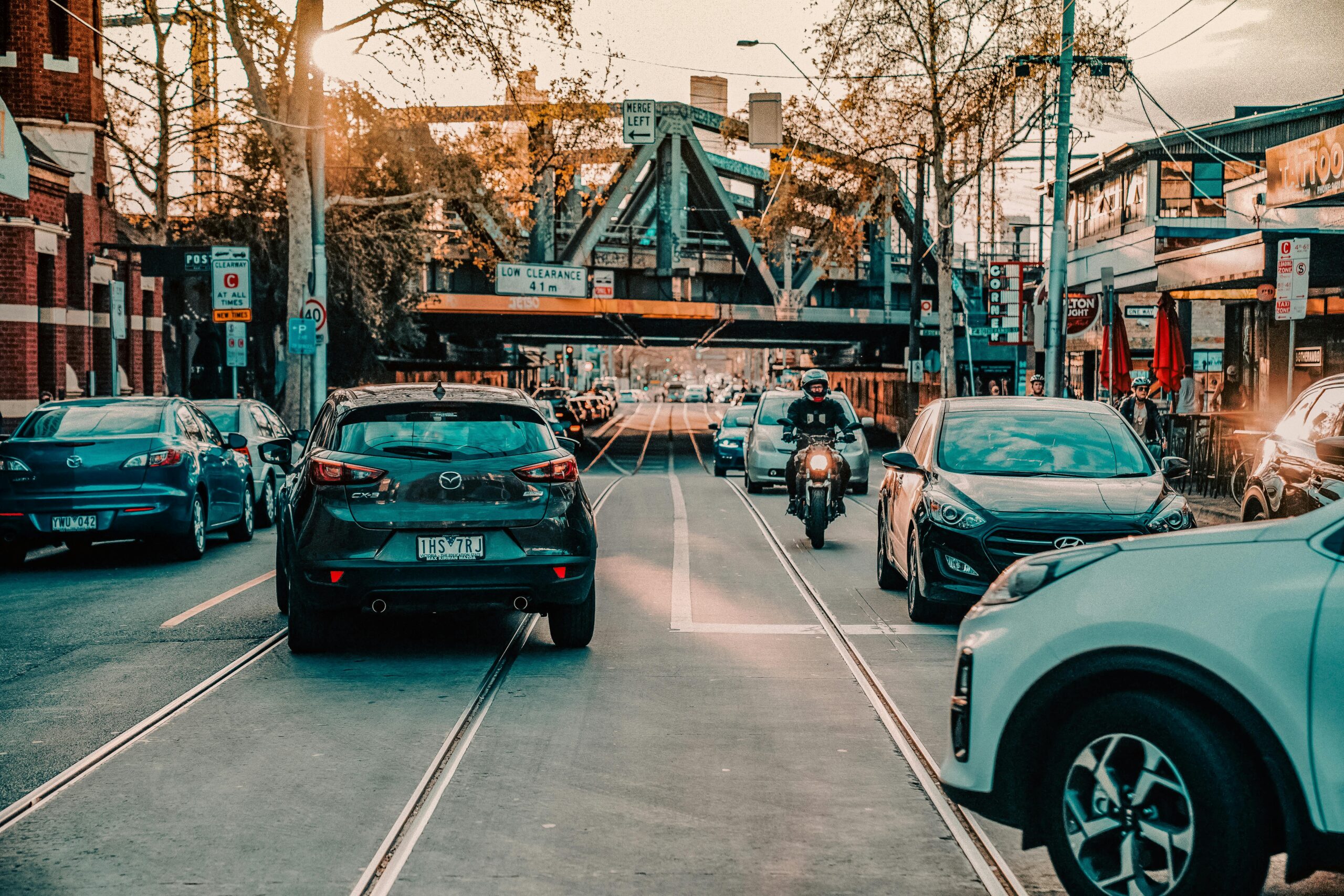 Bustling city street with cars and a motorcycle under a truss bridge during daytime.